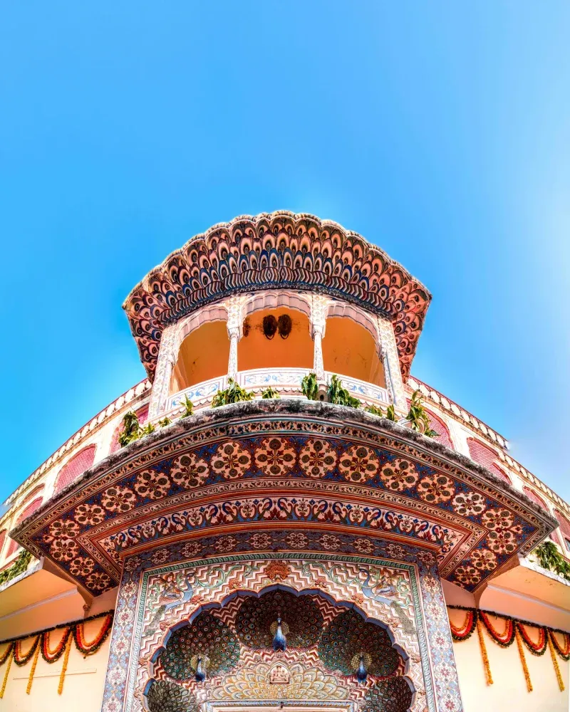 A peacock-frescoed gate at the City Palace seen from below, its scalloped canopy and arched balcony dense with painted florals, marigold garlands at the edges.