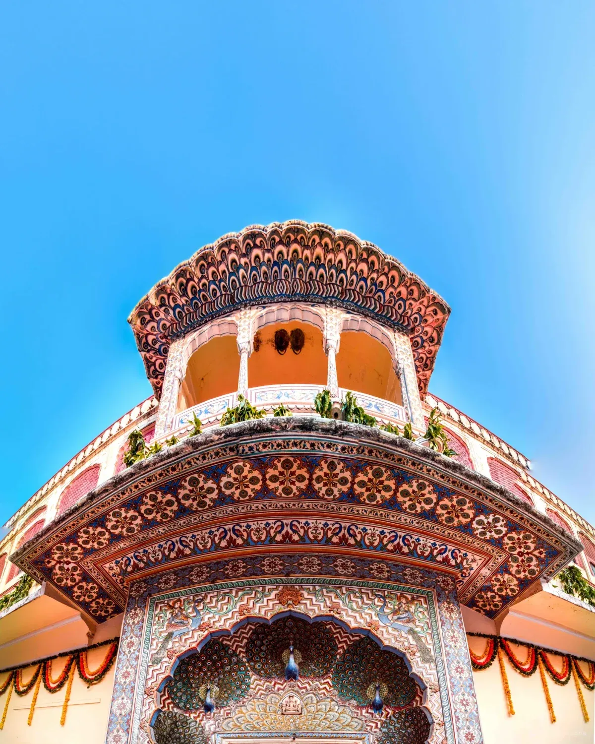 A peacock-frescoed gate at the City Palace seen from below, its scalloped canopy and arched balcony dense with painted florals, marigold garlands at the edges.