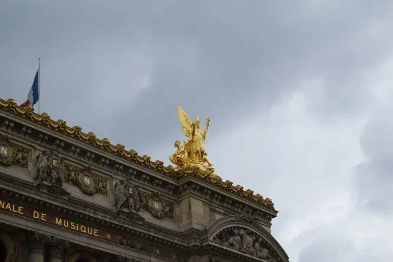 A gilded winged statue on the roofline of the Palais Garnier, a French flag to the left and storm cloud behind.