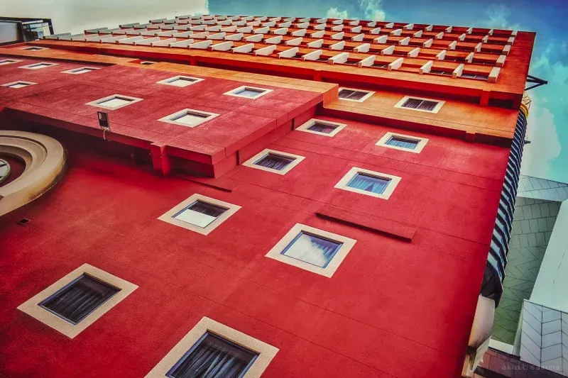A deep red concrete facade tilted steeply upward, square recessed windows in pale surrounds, a slatted canopy against turquoise sky.