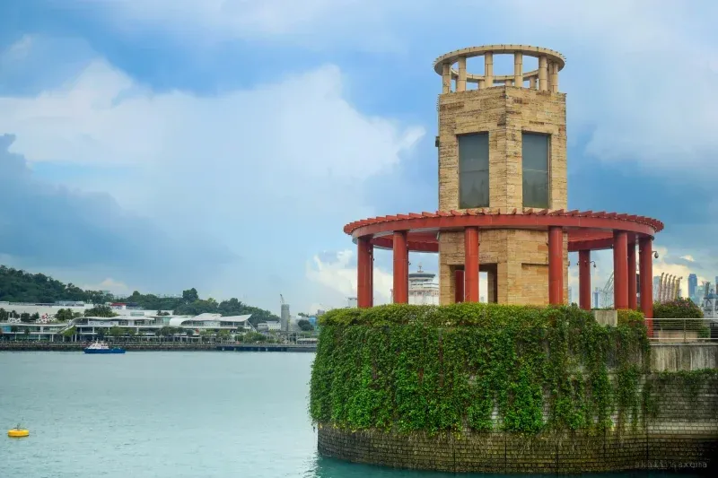A brick tower ringed by a red pergola on a vine-covered island base, harbour buildings and a moored boat across the water.