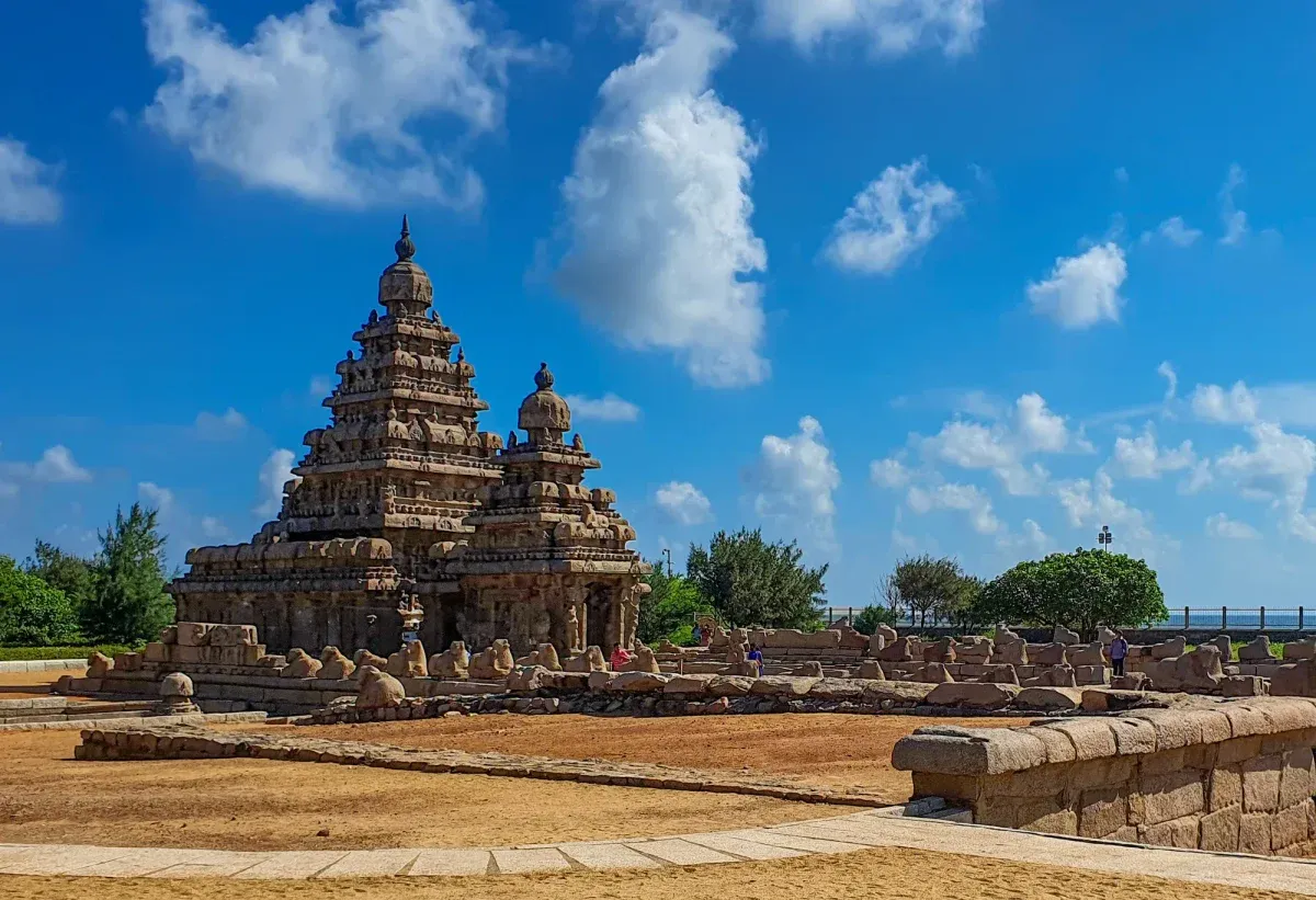 The Shore Temple at Mahabalipuram, its tiered granite tower on a stepped plinth lined with carved bulls, sea horizon to the right under bright cumulus.