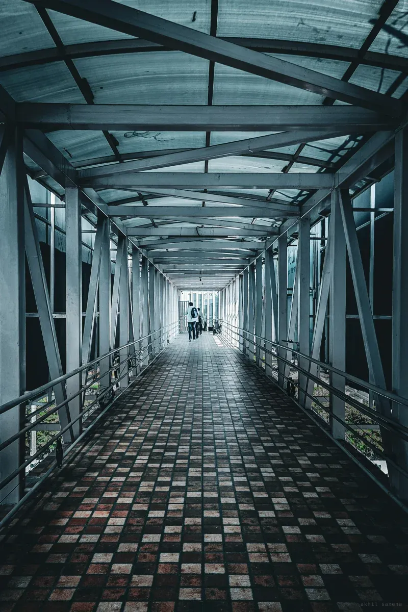 A long covered footbridge of cross-braced steel receding to a bright far end, two figures walking away over red and grey floor tiles.
