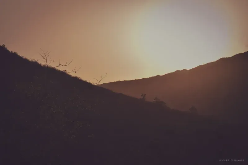 Overlapping hill slopes in sepia haze, a few bare branches on the near ridge, the sun diffused behind.