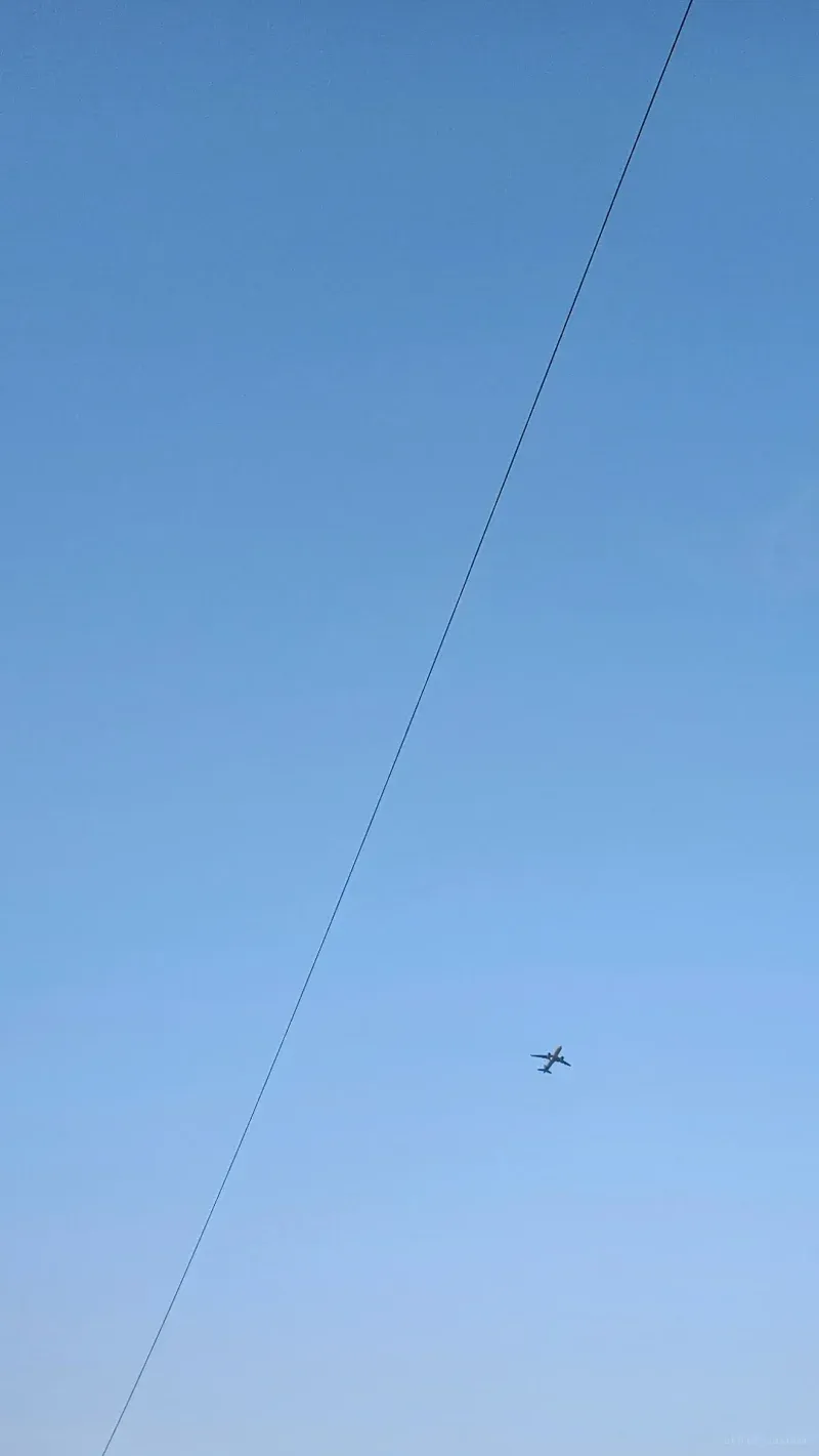 An airliner high in an empty pale blue sky, crossed by a single taut diagonal wire.