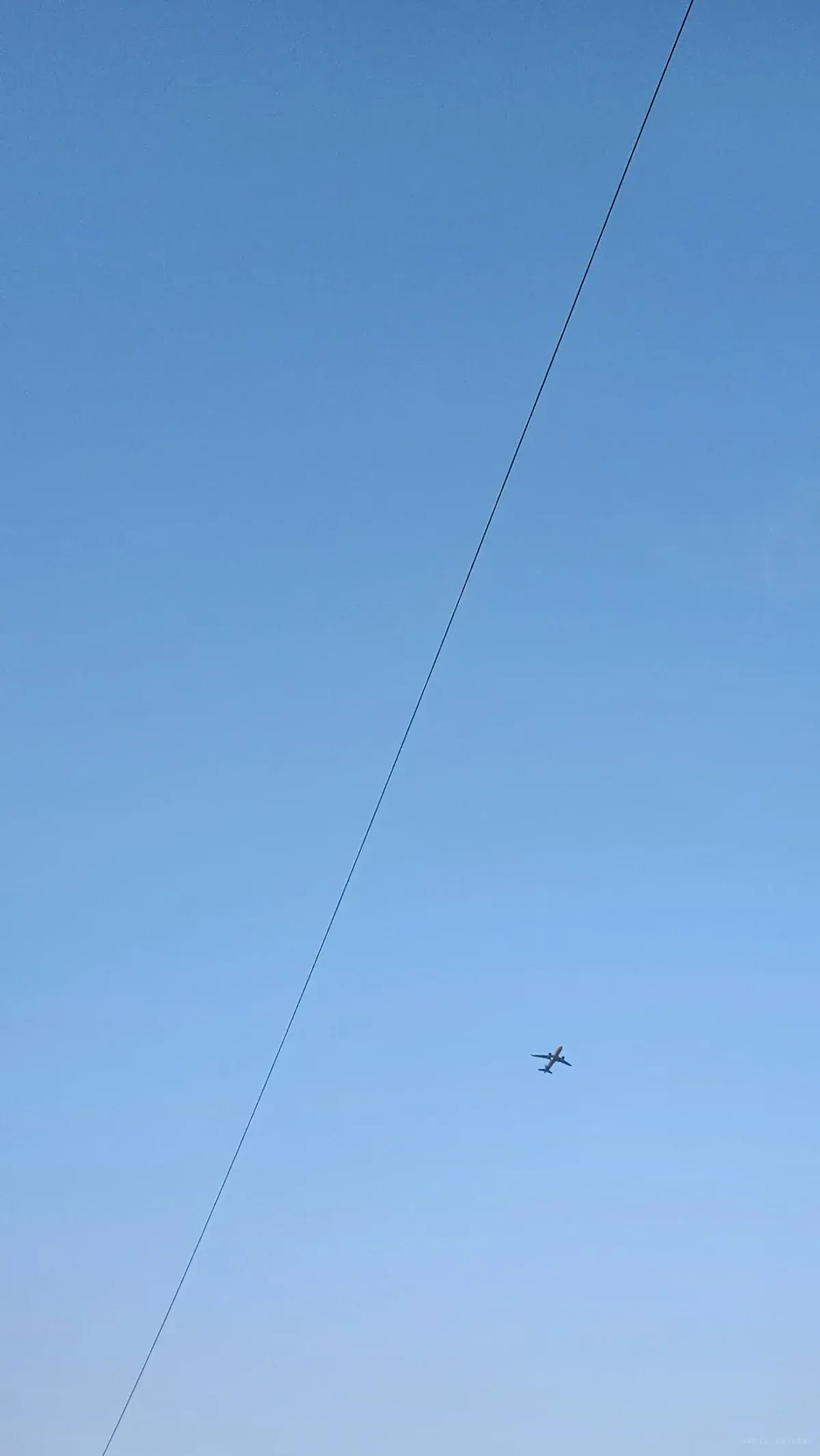 An airliner high in an empty pale blue sky, crossed by a single taut diagonal wire.