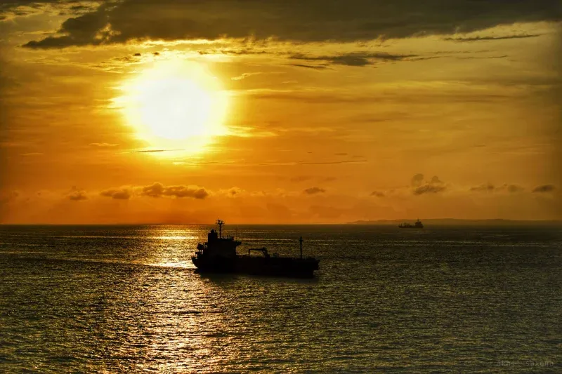A cargo ship in silhouette on open sea beneath a large low sun, a second vessel faint on the horizon.