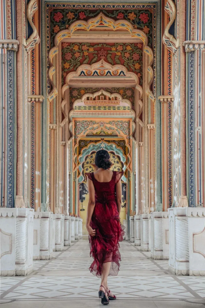 A woman in a deep red ruffled dress walks away from the camera down Patrika Gate's corridor of receding painted archways, each frescoed in florals and teal.