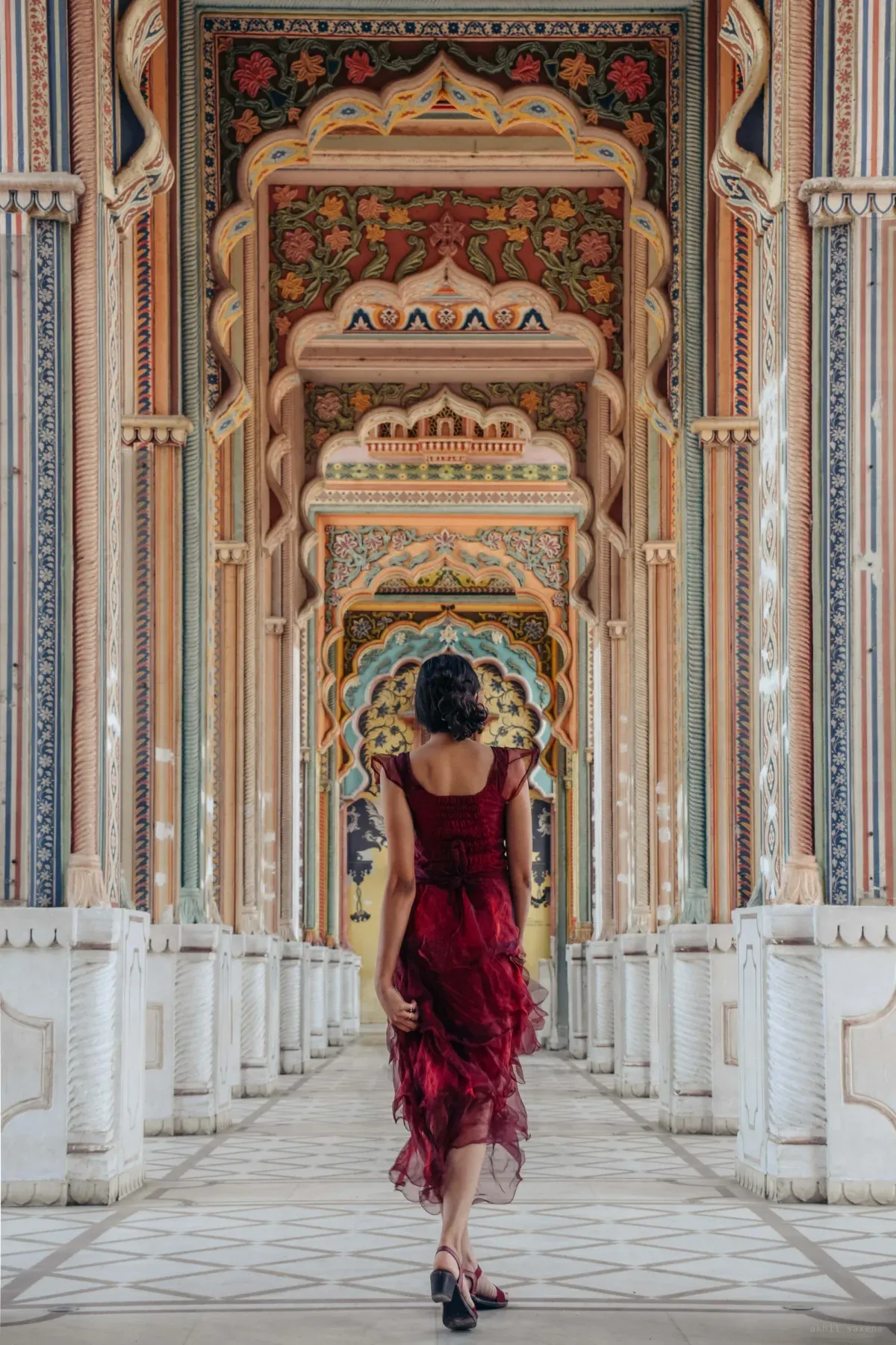 A woman in a deep red ruffled dress walks away from the camera down Patrika Gate's corridor of receding painted archways, each frescoed in florals and teal.