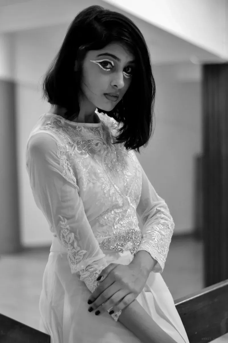 Black-and-white portrait of a woman with a chin-length bob and graphic winged eyeliner, in a sheer embroidered white dress, leaning on a rail.