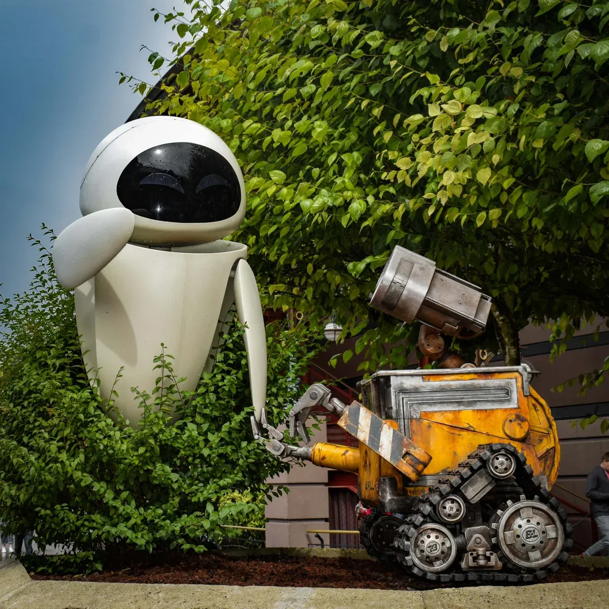 Life-size WALL-E and EVE figures in a planted bed, the white egg-shaped robot leaning toward the rusted tracked one.