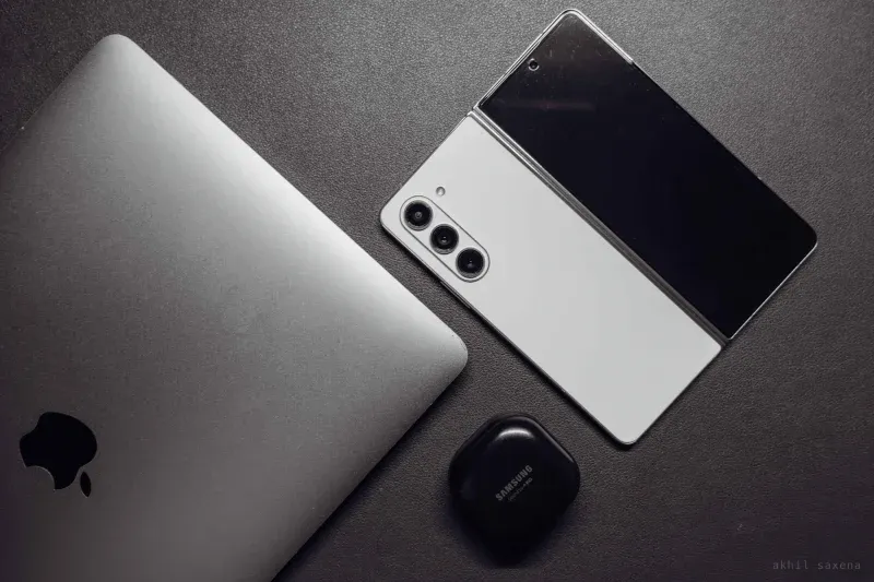 A closed silver laptop, a part-folded phone standing on its hinge, and a small earbuds case, arranged on dark textured slate.
